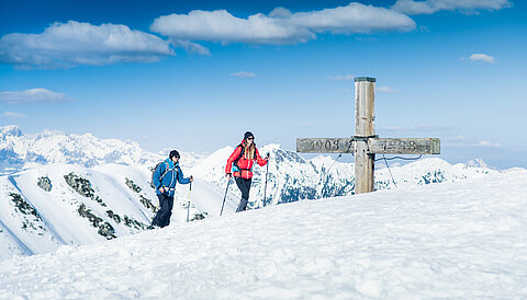 Two ski tourers at the summit cross with panoramic views of the snow-covered Alps in Obertauern