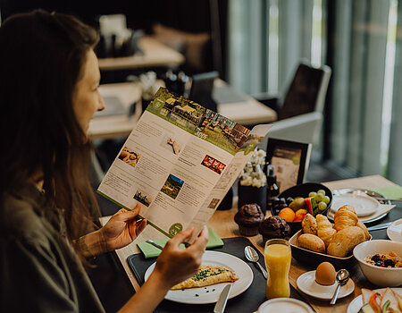 A woman reads a brochure about wellness and leisure activities during breakfast at the Lürzerhof Gourmet & Wellness Hotel.