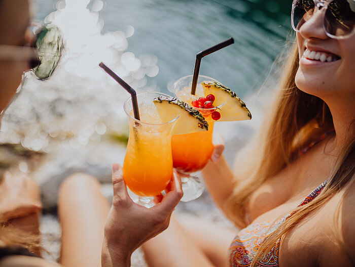 Two women toast each other with cocktails at the mountain lake pool at the Lürzerhof.