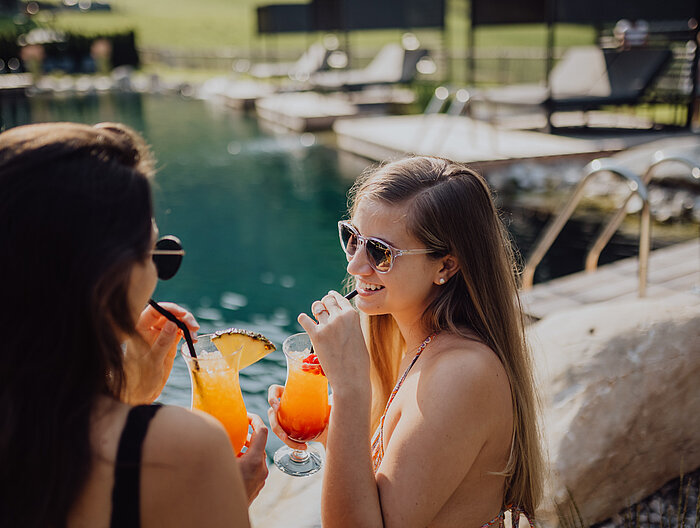 Two women toast each other with cocktails at the mountain lake pool at the Lürzerhof.